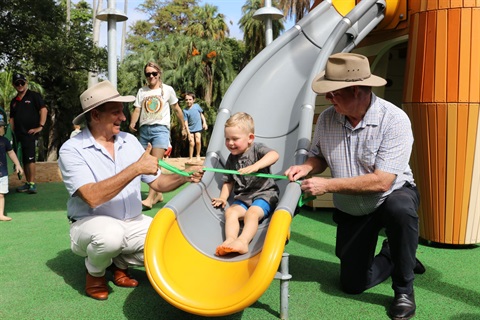 Mayor Tony Williams and MP Barry O'Rourke hold a crepe paper ribbon over a slide as a little boy slides down to officially open the playground
