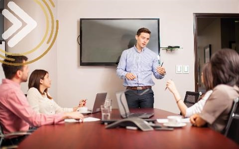 Man standing in front of a projector screen talking to a group of people seated around a board table